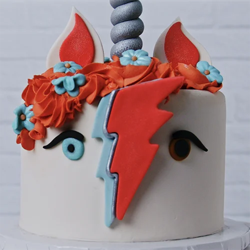 What to make - cake Brightly decorated unicorn-themed birthday cake with colorful fondant, floral accents, and lightning bolt details, perfect for cake decorating supplies and cake decorating tools from Over the Top Cake Supplies.