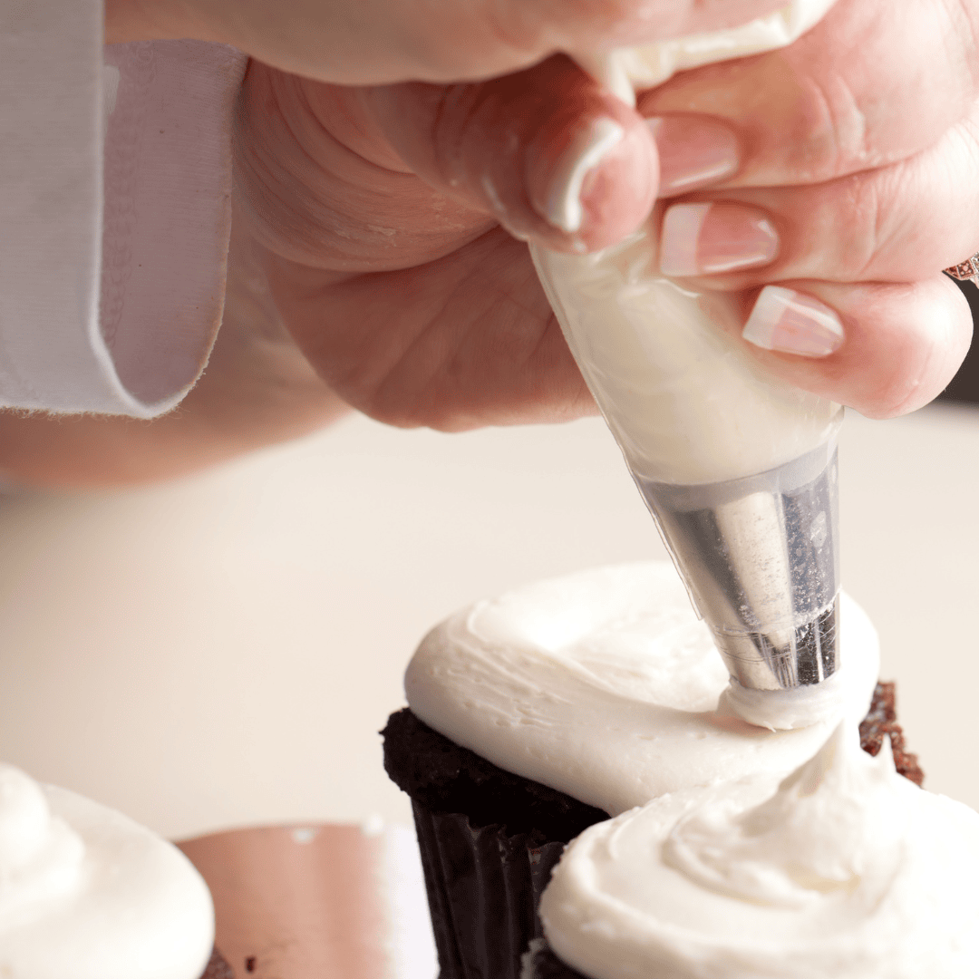 Filling and decorating a chocolate cupcake with white frosting using a piping bag and metal nozzle at Over the Top Cake Supplies.