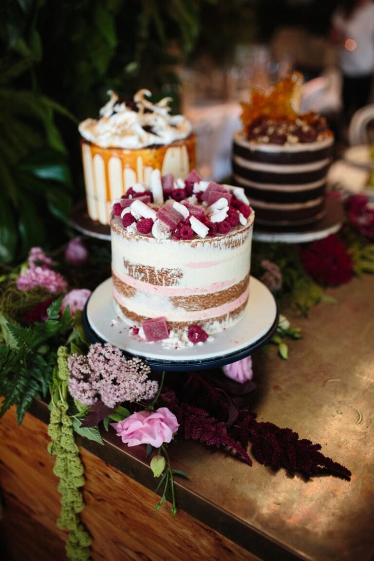 728b63a96f1a8e916bae95a8c7aa06b5 Delicious semi-naked cake with fresh berries and chocolate chunks, perfect for special occasions, on a decorative cake stand surrounded by lush flowers.