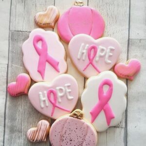 Pink ribbon breast cancer awareness cookies with hope messages and heart-shaped treats, decorated with pink and white icing, are displayed on a rustic wooden surface.