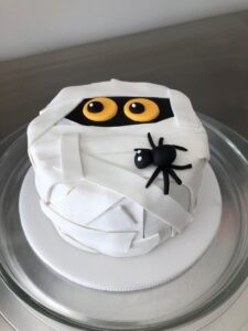 Decorated mummy cake with black spider and yellow eyes, perfect for Halloween celebrations, made with cake decorating supplies from Over the Top Cake Supplies.