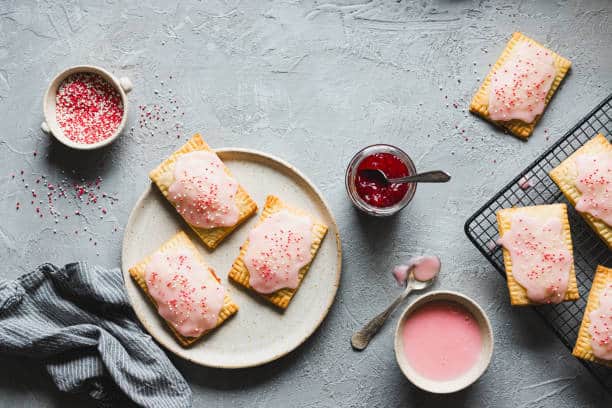 poptarts Homemade fruit jam filled trats on plate with the ingredients on grey table. Table top view of a sweet tarts with strawberry jam, sugar sprinkles and cream.
