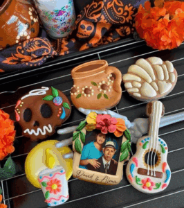 Skull and traditional Mexican sugar skull cookies, decorated with colorful icing, along with a personalized photo cookie, all displayed on a wire rack for Dia de los Muertos celebration.