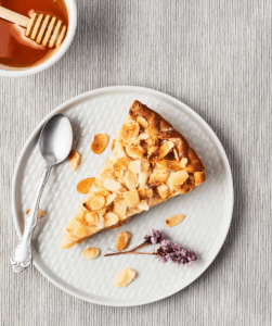Rich almond honey cake slice on white plate with scattered almonds and a purple flower, perfect for cake decorating and baking supplies.