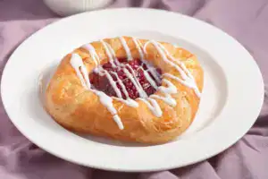 Fresh cherry Danish pastry with white icing drizzle on white plate, showcasing delicious bakery treats from Over the Top Cake Supplies.