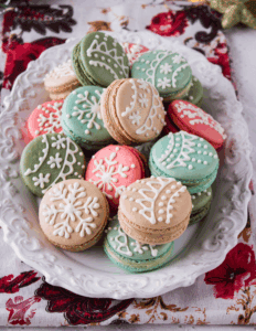 Colorful Christmas macarons decorated with white icing on a decorative white platter, ideal for holiday baking supplies and cake decorating.