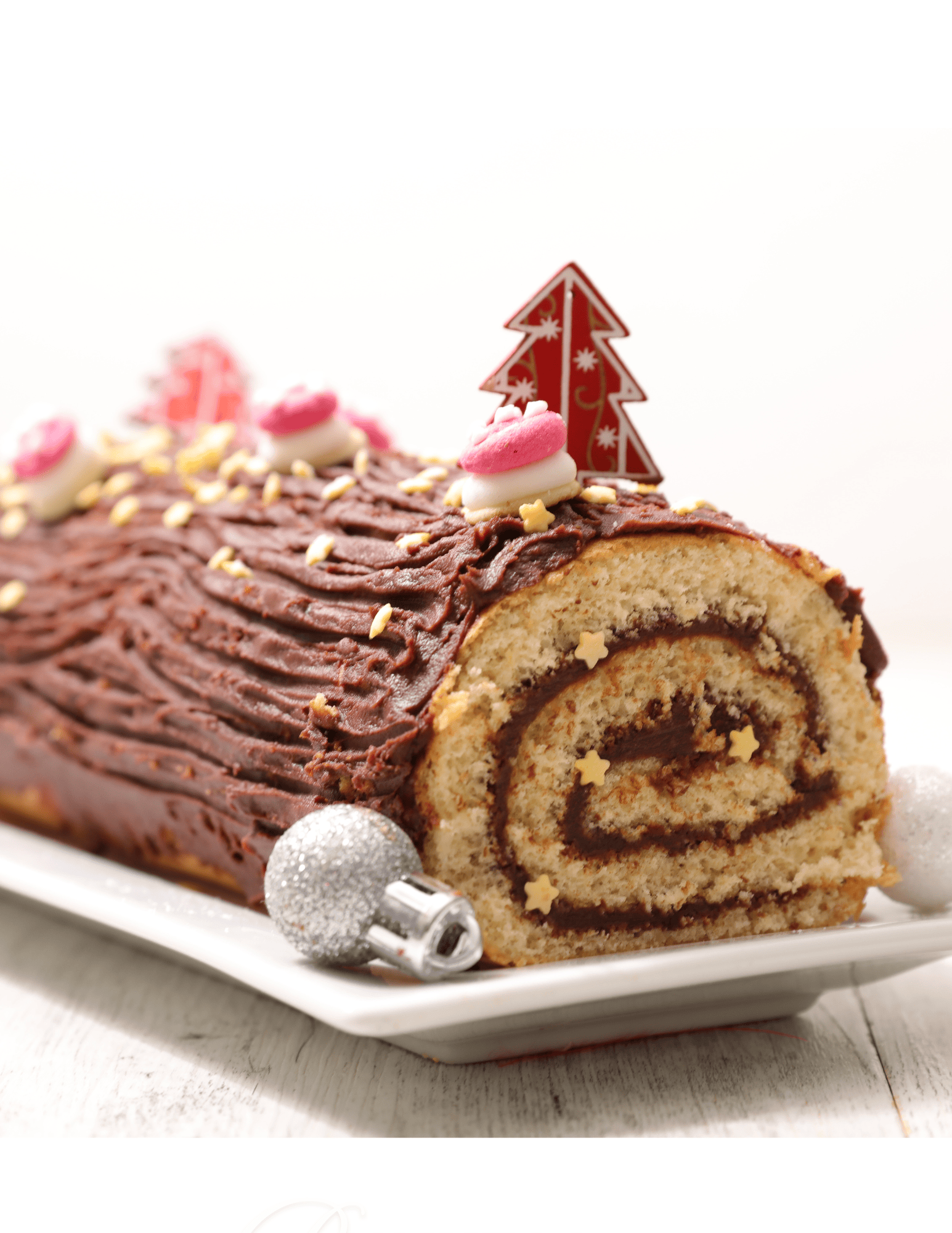 Decorated Christmas yule log cake with chocolate frosting, pink and white fondant accents, edible star sprinkles, and holiday-themed cake topper, on a white serving platter.