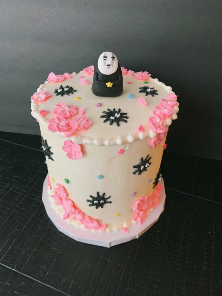spirited away no face cake Decorative birthday cake with pink flower frosting and black snowflake-shaped accents, topped with a unique character cake topper, designed for celebrations.