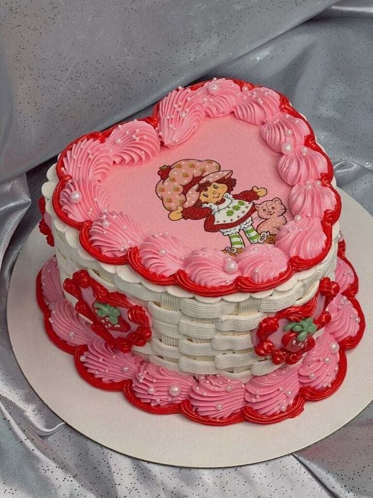 Colorful heart-shaped cake with pink and red frosting, featuring a whimsical cartoon girl with a strawberry, decorated with pearl accents and piped frosting details.