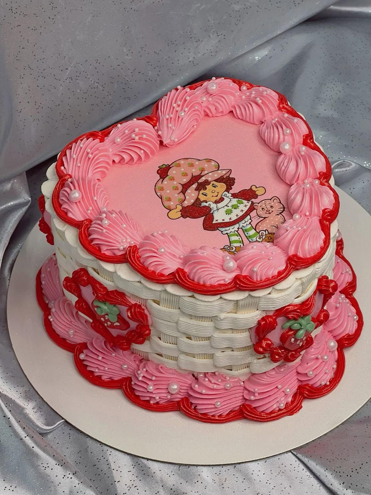 Colorful heart-shaped cake with pink and red frosting, featuring a whimsical cartoon girl with a strawberry, decorated with pearl accents and piped frosting details.