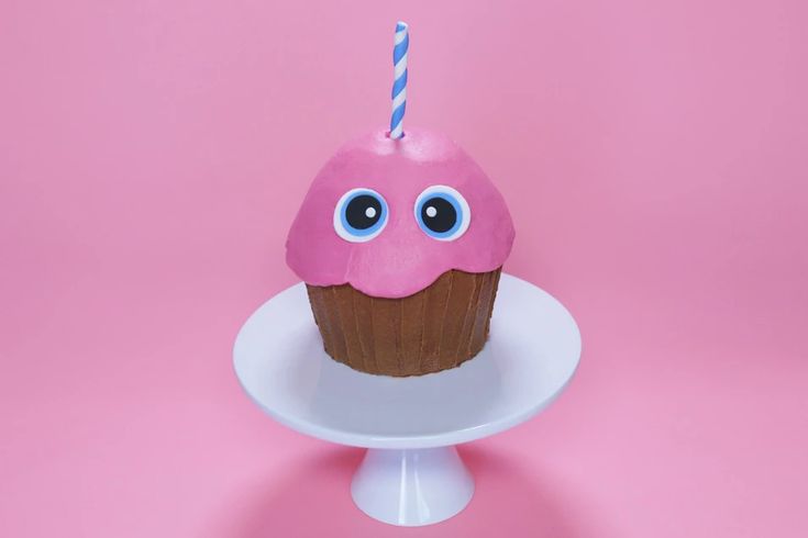 Adorable themed cupcake decorated with pink frosting, cartoon-like cookie eyes, and a blue and white striped candle on a white cake stand against a pink background.