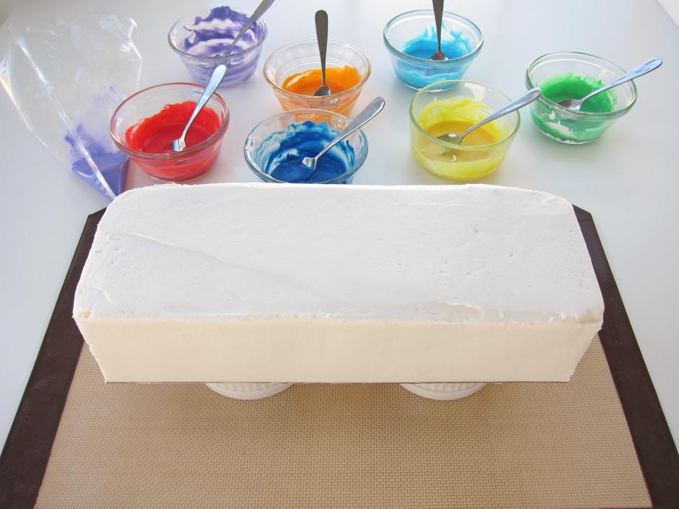 Assorted vibrant cake icing colors in small bowls with spoons, ready for cake decorating. Large white cake base on a baking mat, perfect for creating colorful cake designs.