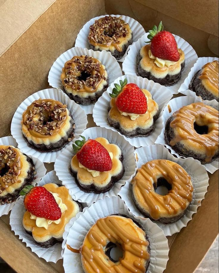 Assorted mini cheesecakes topped with strawberries, caramel drizzle, and crushed nuts in a bakery box, perfect for dessert or party treats.