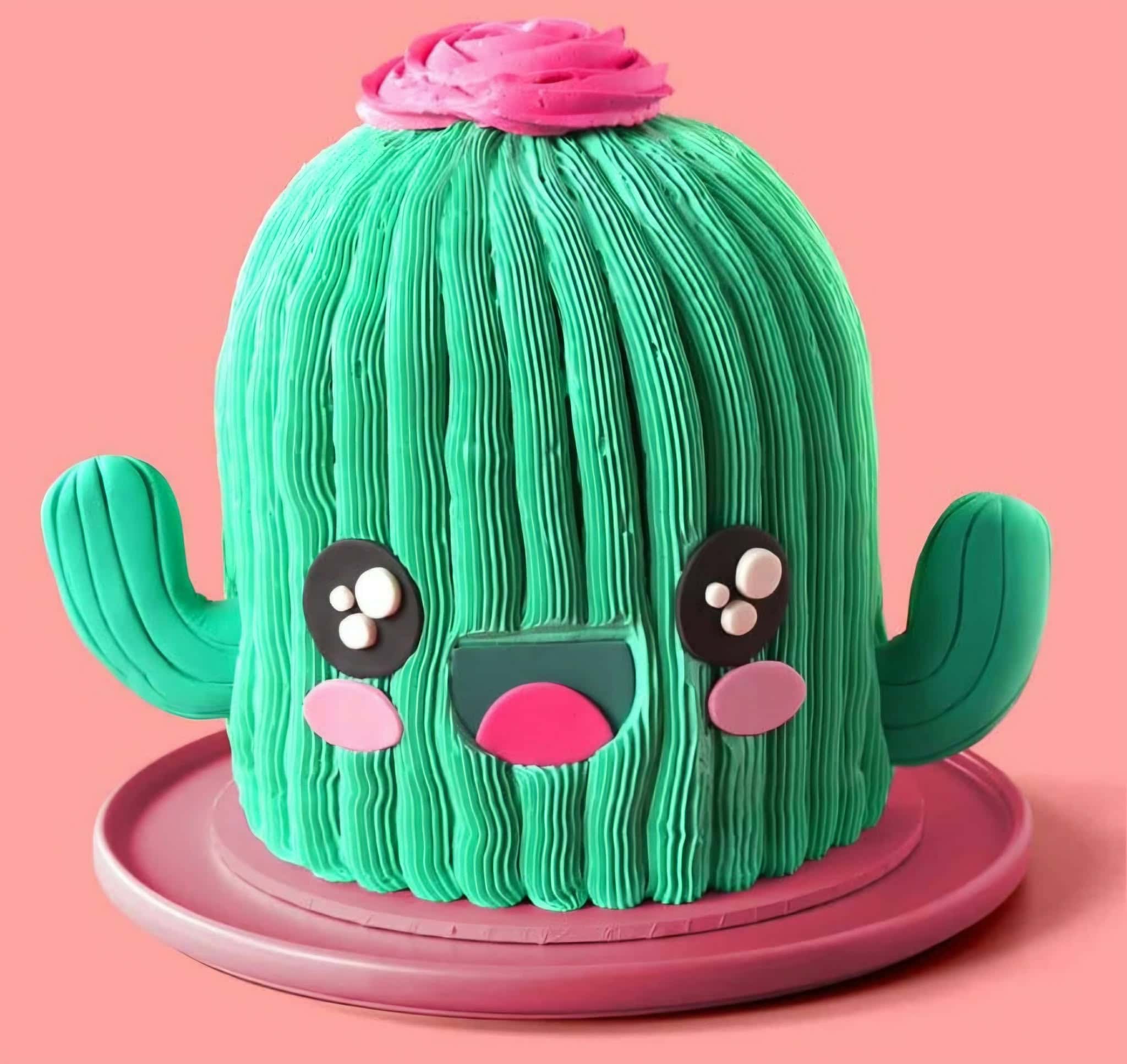 Colorful cactus-shaped cake with a cute face, pink top, and green arms, perfect for celebrations. Made with fondant and decorative details, displayed on a pink plate.