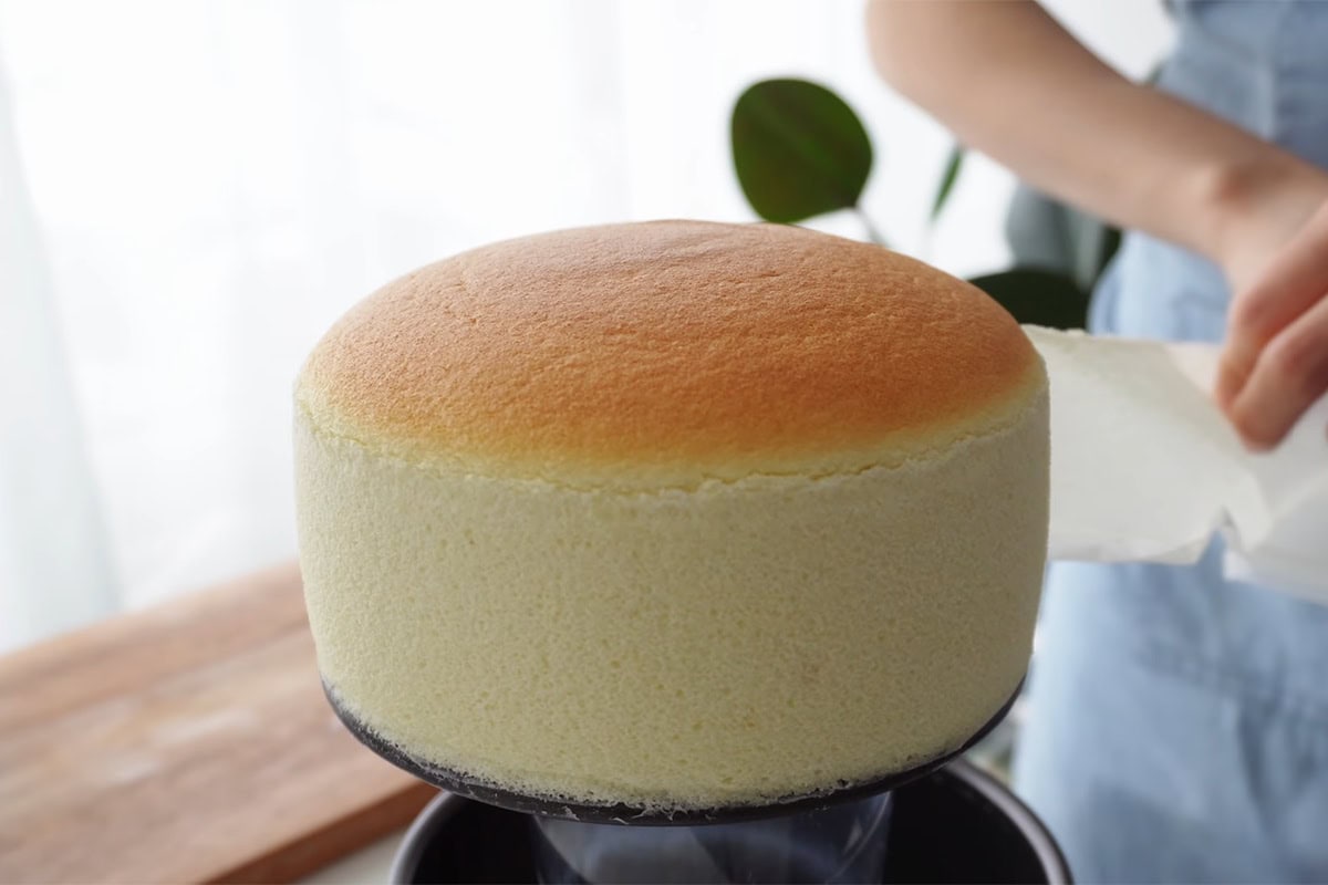Freshly baked round sponge cake on a cake turntable, ready for decorating, with a person in the background preparing to add icing or fondant.