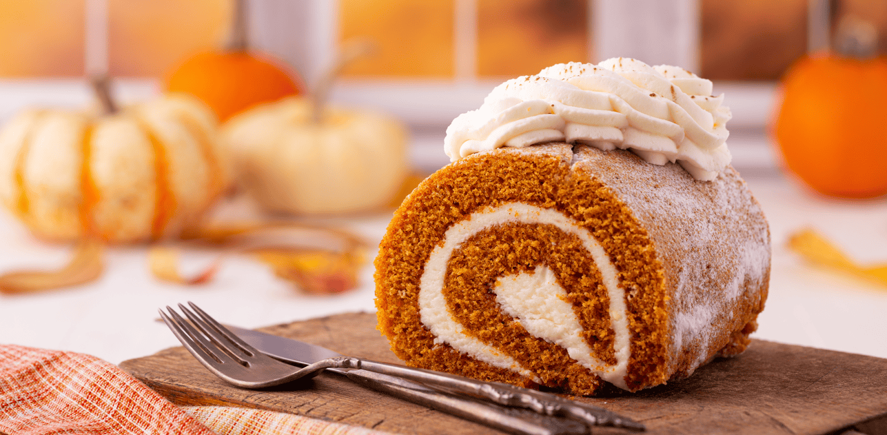 Pumpkin roll cake with cream cheese frosting on a wooden board, surrounded by pumpkins and autumn leaves, perfect for fall desserts and cake decorating supplies.