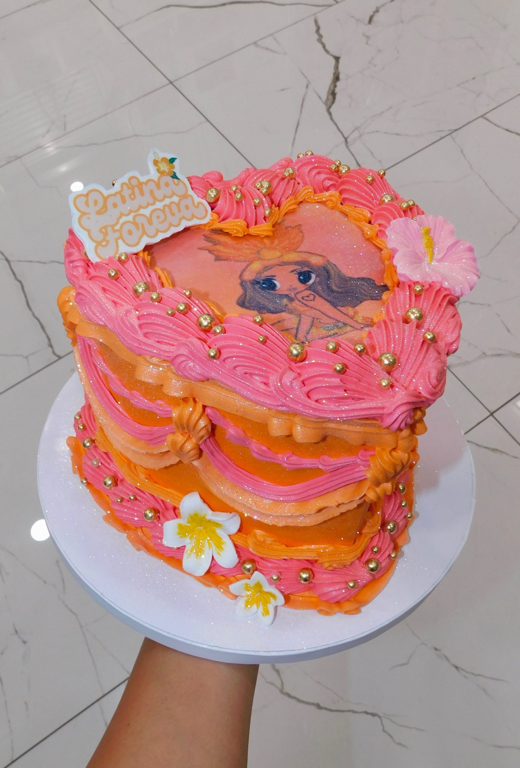 Colorful heart-shaped cake with pink and orange frosting, featuring a printed edible image of Karol G, decorated with gold pearls and flowers for a Latina Foreva themed celebration.