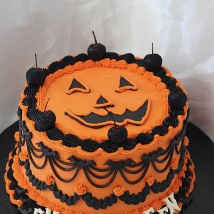 Orange and black Halloween-themed cake with a jack-o'-lantern face, decorated with cherries and intricate icing patterns, perfect for Halloween celebrations and birthday parties.