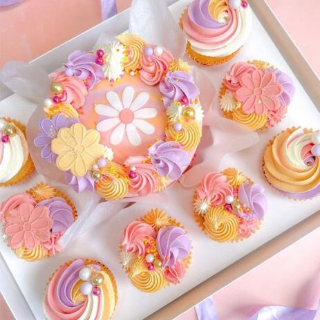 Delightful pastel floral and swirl cupcakes arranged in a spring-themed bento box, featuring pink, purple, and yellow hues with pearl accents and edible flowers, ideal for spring parties.