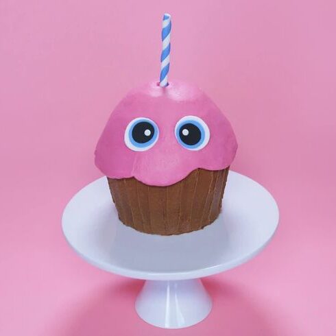 Adorable themed cupcake decorated with pink frosting, cartoon-like cookie eyes, and a blue and white striped candle on a white cake stand against a pink background.