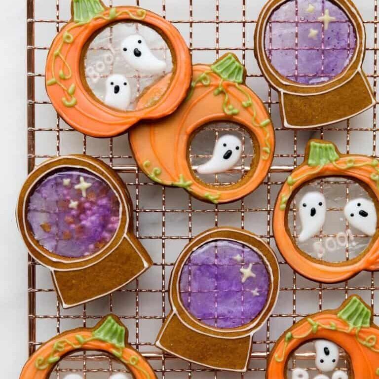 Colorful Halloween ghost and pumpkin cookies with spooky purple coffee cups, decorated with vibrant icing and themed design, displayed on a wire cooling rack. Perfect for Halloween cake decorating and party treats.