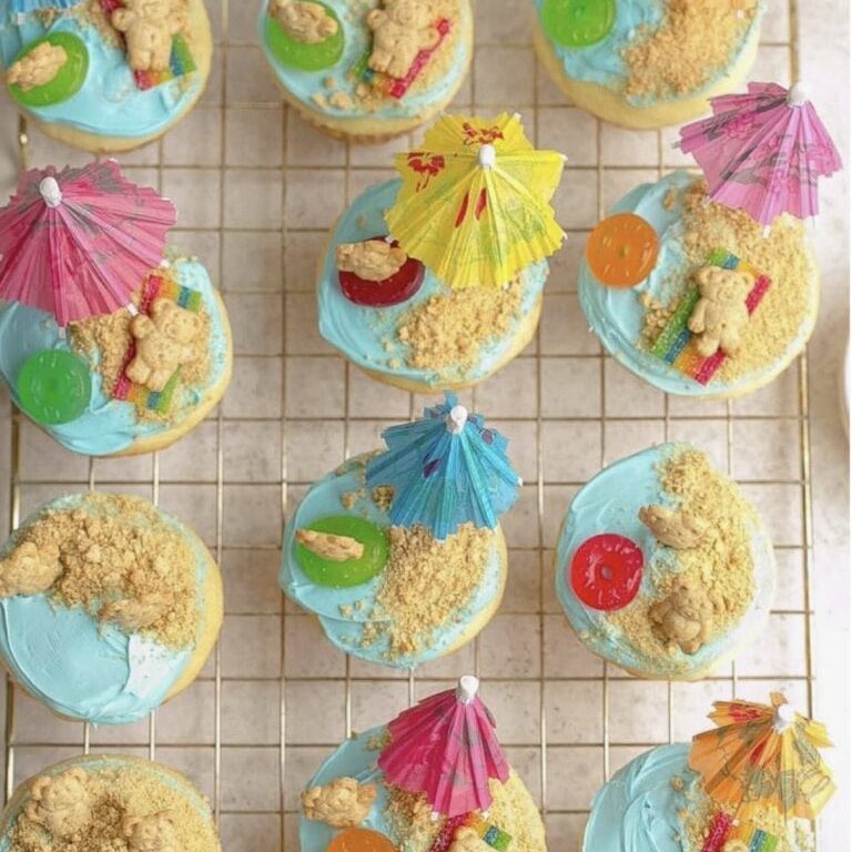 Cupcakes with blue frosting, gummy candies, teddy bear cookies, and colorful umbrellas for a tropical themed celebration, displayed on a wire cooling rack.